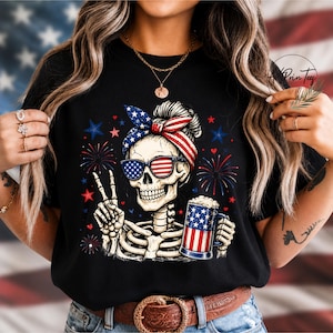 May include: Black t-shirt featuring a skeleton wearing sunglasses and a headband with an American flag design. The skeleton is holding a beer mug with an American flag design and making a peace sign. Fireworks and stars are in the background.