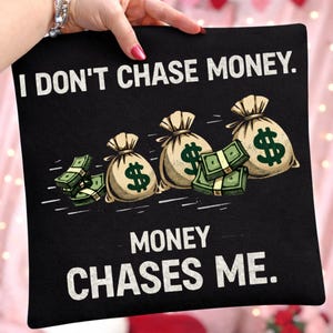 May include: Black pillow with the text "I DON'T CHASE MONEY. MONEY CHASES ME." The design includes illustrations of money bags with dollar signs and stacks of cash. The pillow is held by a person.