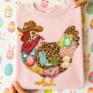 May include: Pink sweatshirt featuring a patchwork chicken design. The chicken wears a cowboy hat and bandana, with patches of leopard print, floral patterns, and Easter-themed elements like eggs and a bunny. The design incorporates a cowboy boot and cactus.