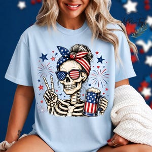 May include: Light blue t-shirt featuring a skeleton wearing sunglasses and a headband with an American flag design. The skeleton is holding a beer mug with an American flag design and making a peace sign. Fireworks and stars are in the background.