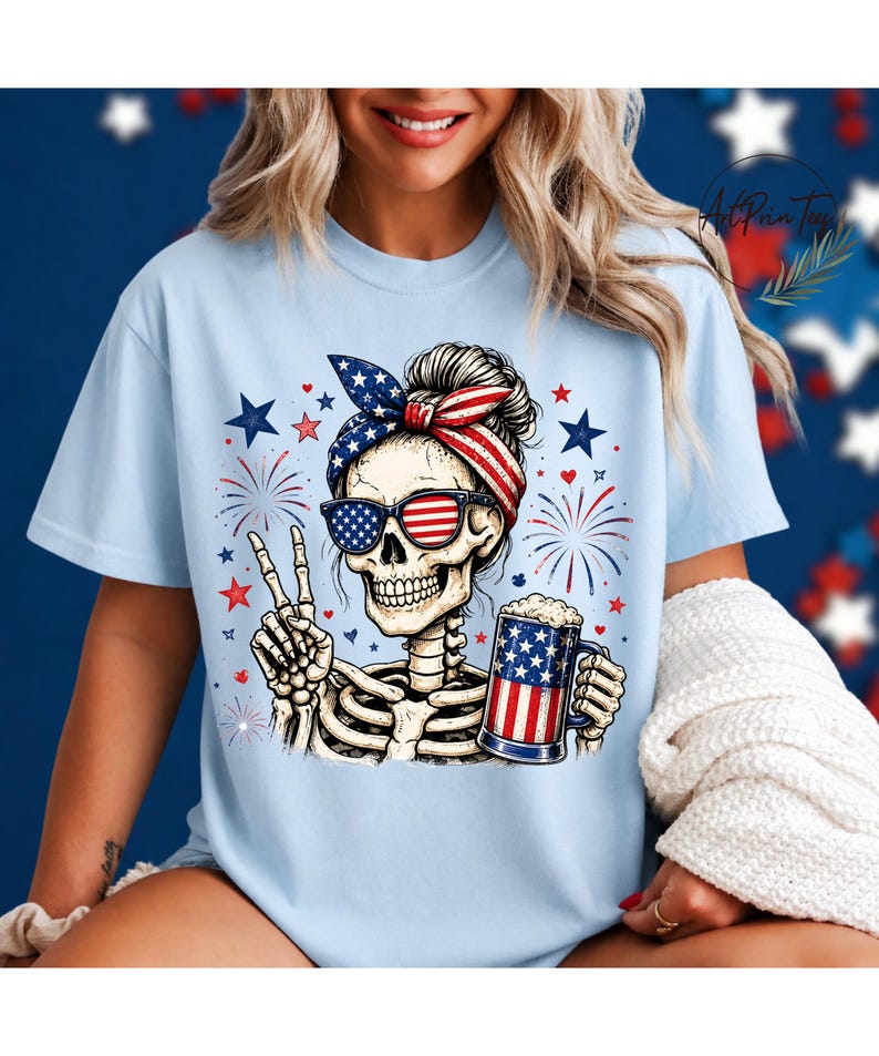 May include: Light blue t-shirt featuring a skeleton wearing sunglasses and a headband with an American flag design. The skeleton is holding a beer mug with an American flag design and making a peace sign. Fireworks and stars are in the background.