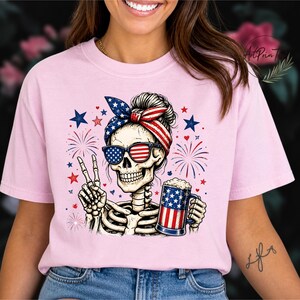 May include: Pink t-shirt featuring a skeleton with sunglasses and a headband in the colors of the American flag. The skeleton is holding a beer mug with a flag design and making a peace sign. Fireworks and stars complete the patriotic design.