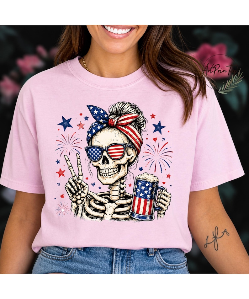 May include: Pink t-shirt featuring a skeleton with sunglasses and a headband in the colors of the American flag. The skeleton is holding a beer mug with a flag design and making a peace sign. Fireworks and stars complete the patriotic design.