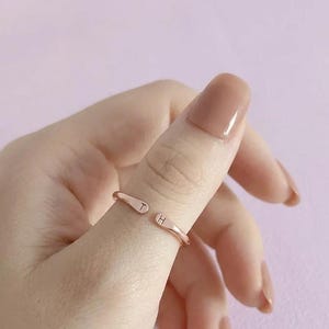 May include: A rose gold-toned open ring featuring two flat, rounded ends. Each end is engraved with a single uppercase letter, with the letter T on one side and the letter H on the other. The band has a smooth, polished finish and a simple design.