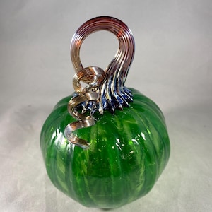 May include: A handcrafted glass pumpkin with a vibrant green body and a metallic silver and copper stem. The pumpkin's surface has a glossy finish, reflecting light. The stem is a looped design, adding a decorative touch to this fall-themed item.