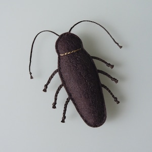 May include: A handmade, dark brown felt insect with six braided legs and two long antennae. The insect has a gold-colored band around its body. The toy is designed to resemble a cockroach.