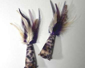 May include: Two cat toys with a cone-shaped base and feather top. The base has a spotted pattern in shades of brown and purple. The feathers are purple, cream, and brown. The toys are designed for cats.