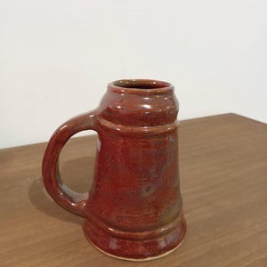 Peut inclure: Une chope en céramique rouge faite à la main avec une anse. La chope a une forme cylindrique avec une base légèrement évasée et une anse arrondie. L'émail a une finition brillante avec des variations de couleur.