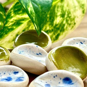 Peut inclure: Un ensemble de petits plats en céramique en forme de galets. Certains sont vernis d'un vert olive brillant, tandis que d'autres ont une base blanche avec des mouchetures bleues. Les plats sont disposés en grappe, avec des feuilles vertes et jaunes en arrière-plan.