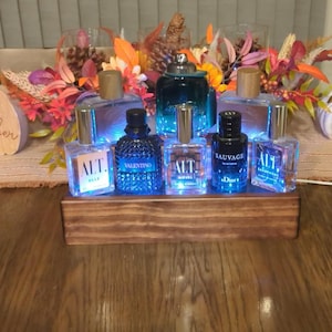 May include: A wooden display stand holding several perfume bottles. The bottles are of different shapes and sizes, made of clear and coloured glass. Some bottles have gold or silver caps. The stand is lit with blue lights, creating an attractive display.