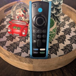 May include: Black remote control with a light blue protective case. The remote has several buttons, including a circular navigation pad and buttons for Disney+ and Hulu. The remote is on a wooden tray with a Christmas tree pattern.