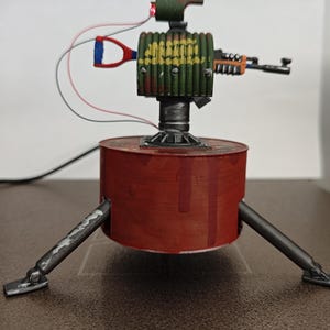 Könnte beinhalten: Ein handgefertigter, miniaturisierter Roboterturm mit roter Basis und drei Beinen. Der Turm hat einen grün-gelben zylindrischen Körper, eine kleine Waffe und ein rotes Licht oben. Drähte erstrecken sich von oben und der Seite.