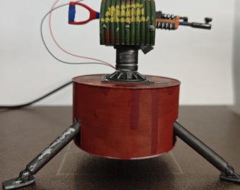 May include: A handmade, miniature, robotic turret with a red base and three legs. The turret has a green and yellow cylindrical body, a small gun, and a red light on top. Wires extend from the top and side.
