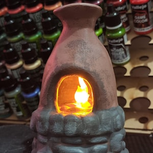 May include: A small, brown ceramic fireplace with a glowing orange light inside, resembling a flame. The base is a darker gray, stone-like texture. The background shows various paint bottles.