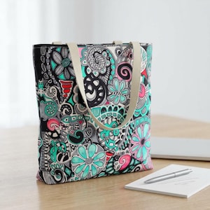 Peut inclure: Un sac fourre-tout coloré avec un motif floral noir, turquoise, rose et gris. Le sac a des anses beiges et est fait d'un tissu robuste. Le design présente des fleurs stylisées et des formes abstraites.