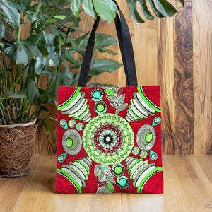 Könnte beinhalten: Rote Tragetasche mit einem symmetrischen Design in Grün, Weiß und Türkis. Das Muster zeigt ein zentrales Mandala-ähnliches Motiv und sich wiederholende geometrische Formen. Schwarze Griffe kontrastieren mit dem farbenfrohen Druck.
