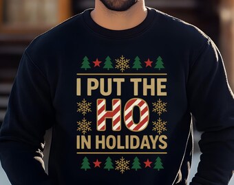 May include: Black sweatshirt with the text "I PUT THE HO IN HOLIDAYS" in gold and red. The design includes Christmas trees, stars, and snowflakes. The person in the image is wearing a black beanie.