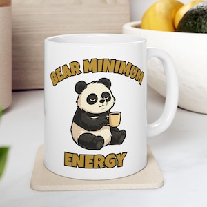 Puede incluir: Taza de cerámica blanca con un oso panda de dibujos animados sosteniendo una pequeña taza amarilla. El texto "BEAR MINIMUM ENERGY" está impreso en letras doradas encima y debajo de la ilustración del panda. La taza está sobre un posavasos beige.