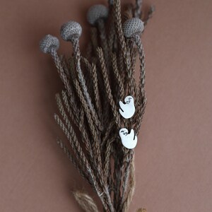 May include: Two white sloth earrings displayed on a dried floral arrangement. The earrings feature a simple sloth design. The background is a plain brown, providing a neutral backdrop for the earrings and the floral arrangement.
