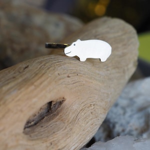 Pode incluir: Anel de prata com um pequeno pingente de hipopótamo branco. O hipopótamo tem um design simples, tipo desenho animado, com um rosto sorridente. O anel está sobre um fundo de madeira natural.