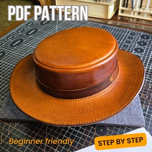 May include: A handcrafted, tan leather hat with a dark brown band and brim. The hat has a textured surface and is labeled "PDF PATTERN", "Beginner friendly", and "STEP BY STEP". The hat appears to be a DIY project.