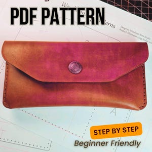 May include: A handmade leather wallet in shades of brown and pink, secured with a decorative button. The text "PDF PATTERN" is at the top, with "STEP BY STEP" and "Beginner Friendly" at the bottom, indicating a DIY project.