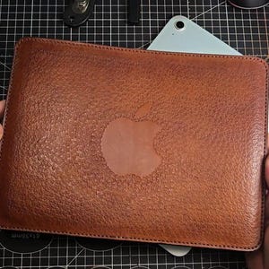 Könnte beinhalten: Eine braune Tablet-Hülle aus Leder mit geprägtem Apfel-Logo. Die Hülle hat eine strukturierte Oberfläche und ist für ein Tablet konzipiert. Der Artikel wird von Händen gehalten.