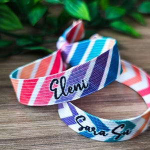 Könnte beinhalten: Zwei bunte elastische Haargummis mit diagonalen Streifen in Rosa-, Orange-, Blau- und Lilatönen. Eines ist verknotet, das andere ist eine Schlaufe. Der Name "Eleni" ist auf einem und "Sara Su" auf dem anderen gestickt.
