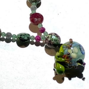 Puede incluir: Un collar con un colgante de rana y una cadena de cuentas. El collar tiene cuentas verdes, rosas y granates. El colgante es una rana verde que abraza una cuenta azul y blanca en forma de globo con un diseño floral.