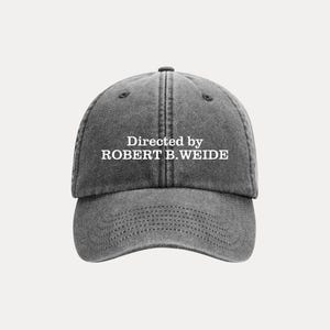 May include: Grey baseball cap featuring the text "Directed by ROBERT B. WEIDE" in white. The cap has a curved peak and a stitched design. The hat is made of a soft material.