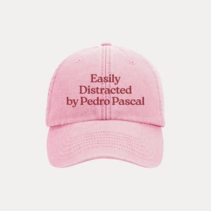 May include: A pink baseball cap with the text "Easily Distracted by Pedro Pascal" in a darker pink font. The cap has a curved brim and an adjustable strap.