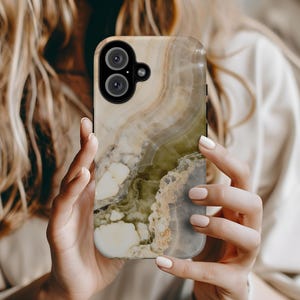 May include: A smartphone case with a marble design in shades of beige, green, and gray. The case has a black camera cutout and is held by a person with light skin. The case has a glossy finish.