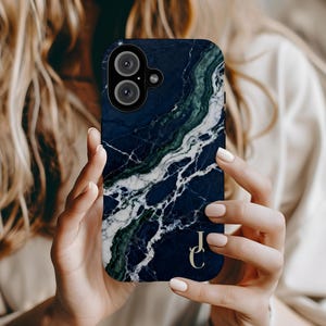 May include: A smartphone case with a dark blue marble design, featuring white and green veining. The case has a camera cutout and gold initials "JC" on the lower right corner. The case is held by a person.