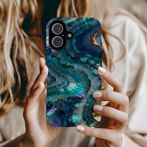 May include: A smartphone case with a swirling, agate-like design in shades of teal, blue, and gold. The case has a glossy finish and a camera cutout. The case is held by a person with light skin.