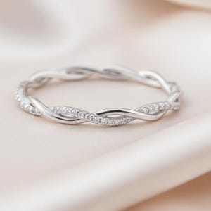 May include: A silver-colored ring with a braided design. The ring is adorned with small, clear gemstones that sparkle. The ring is set against a soft, light-colored background, highlighting its elegant design.