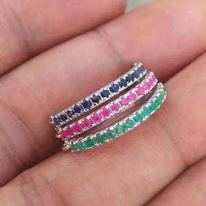 Può includere: Tre anelli d'argento impilati, ciascuno ornato da una fila di piccole pietre preziose. L'anello superiore presenta pietre blu, l'anello centrale ha pietre rosa e l'anello inferiore è verde. Gli anelli sono esposti su uno sfondo neutro.