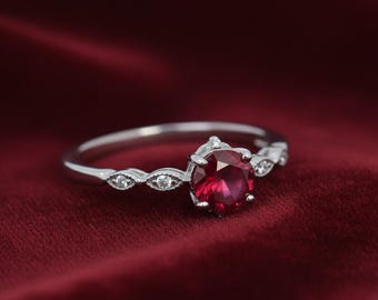 Puede incluir: Un anillo plateado con una gran gema roja redonda como punto focal. La banda presenta delicados detalles decorativos con pequeñas piedras claras. El anillo se encuentra sobre un fondo rojo oscuro texturizado.