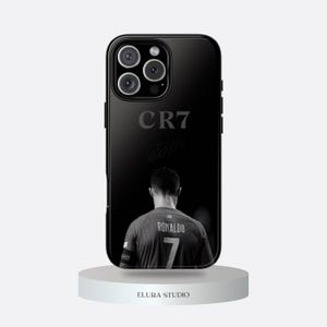 K&ouml;nnte beinhalten: Schwarze Handyh&uuml;lle mit einem Graustufenbild eines Fu&szlig;ballspielers, der "RONALDO" und die Nummer "7" auf dem Trikot zeigt. Die H&uuml;lle zeigt auch "CR7" und eine Unterschrift. Die Handyh&uuml;lle steht auf einem wei&szlig;en Podest.
