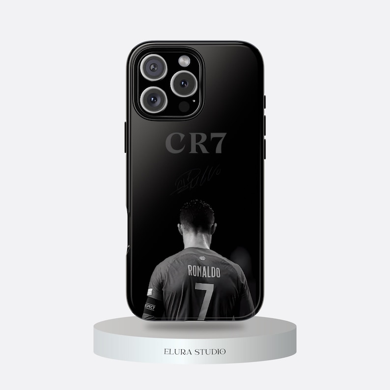 K&ouml;nnte beinhalten: Schwarze Handyh&uuml;lle mit einem Graustufenbild eines Fu&szlig;ballspielers, der "RONALDO" und die Nummer "7" auf dem Trikot zeigt. Die H&uuml;lle zeigt auch "CR7" und eine Unterschrift. Die Handyh&uuml;lle steht auf einem wei&szlig;en Podest.
