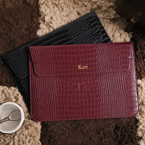 May include: Two leather laptop sleeves with a crocodile texture. One is black, and the other is burgundy with the name "Keri" in gold lettering. The sleeves are on a patterned brown and beige rug.