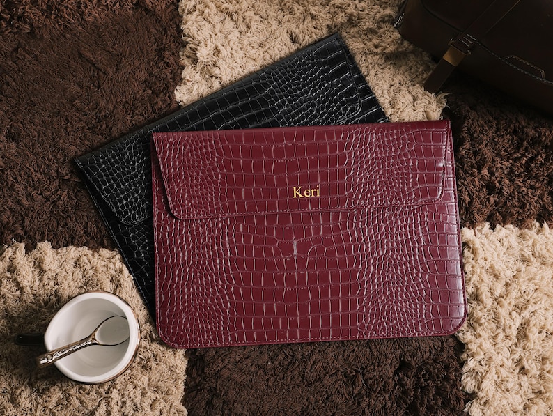 May include: Two leather laptop sleeves with a crocodile texture. One is black, and the other is burgundy with the name "Keri" in gold lettering. The sleeves are on a patterned brown and beige rug.