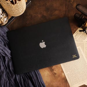 Peut inclure: Un autocollant noir pour ordinateur portable avec un logo Apple argenté centré et les initiales JK dorées en relief dans le coin inférieur droit. La surface est texturée et posée à plat, présentant une finition mate élégante aux bords nets.