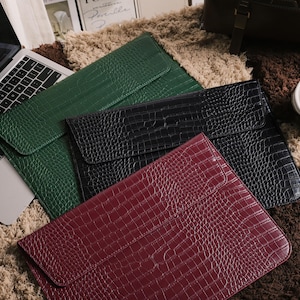 May include: A collection of laptop sleeves in green, black, and burgundy, featuring a crocodile skin texture. The sleeves are arranged on a textured, multi-toned rug, with a laptop visible in the background.