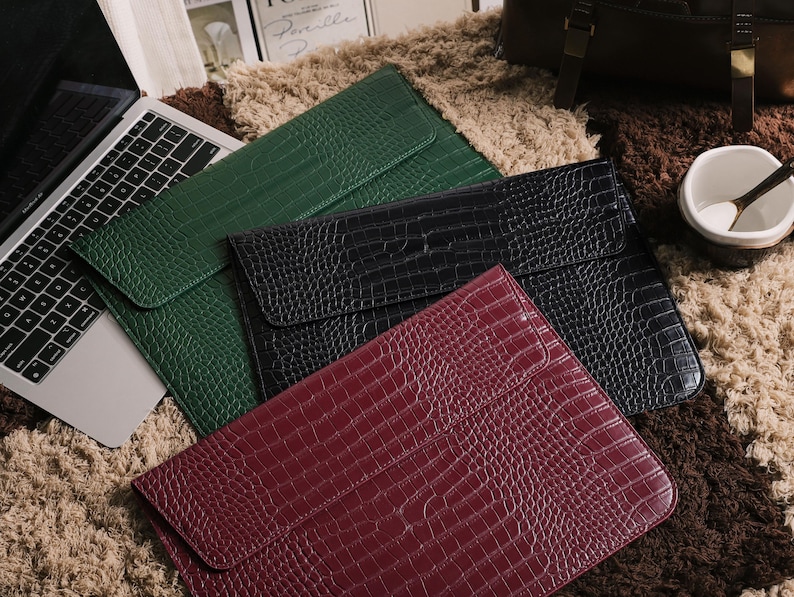 May include: A collection of laptop sleeves in green, black, and burgundy, featuring a crocodile skin texture. The sleeves are arranged on a textured, multi-toned rug, with a laptop visible in the background.
