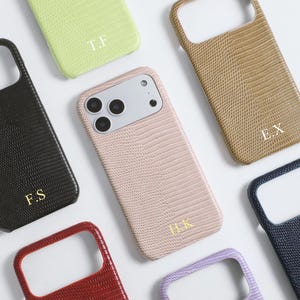 Pode incluir: Uma coleção de capas de telefone personalizadas em várias cores, incluindo preto, verde, castanho, rosa, vermelho e roxo. Cada capa apresenta um padrão texturizado, semelhante à pele de réptil, e iniciais douradas. As capas estão dispostas sobre uma superfície branca.