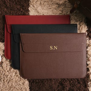 Puede incluir: Tres fundas para portátiles en rojo, negro y marrón, dispuestas sobre una superficie texturizada. La funda marrón tiene las iniciales doradas "S.N" en la parte delantera. Las fundas tienen un diseño sencillo y minimalista con cierre de solapa. La estética general es moderna y funcional.