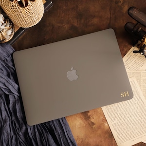 Op de afbeelding: Een matgrijze laptop met een zilverkleurig Apple-logo in het midden van het deksel en de goudkleurige initialen SH in de rechterbenedenhoek. De laptop ligt plat en toont de gladde, effen buitenkant en het strakke ontwerp van de behuizing.