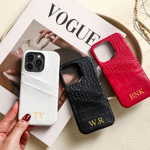 Puede incluir: Tres fundas para teléfono con textura de cocodrilo. Una es blanca con "TY" en dorado, una negra con "W.R" en dorado y una roja con "BNK" en dorado. Las fundas están sobre una revista con la palabra "VOGUE" visible.