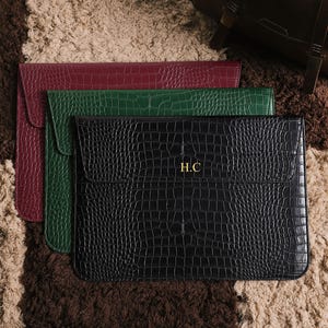 May include: Three leather laptop sleeves in burgundy, green, and black, arranged on a textured rug. The black sleeve features the gold initials "H.C." and a crocodile skin pattern. The sleeves have a flap closure.