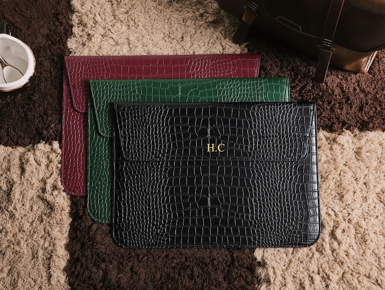 May include: Three leather laptop sleeves in burgundy, green, and black, arranged on a textured rug. The black sleeve features the gold initials "H.C." and a crocodile skin pattern. The sleeves have a flap closure.